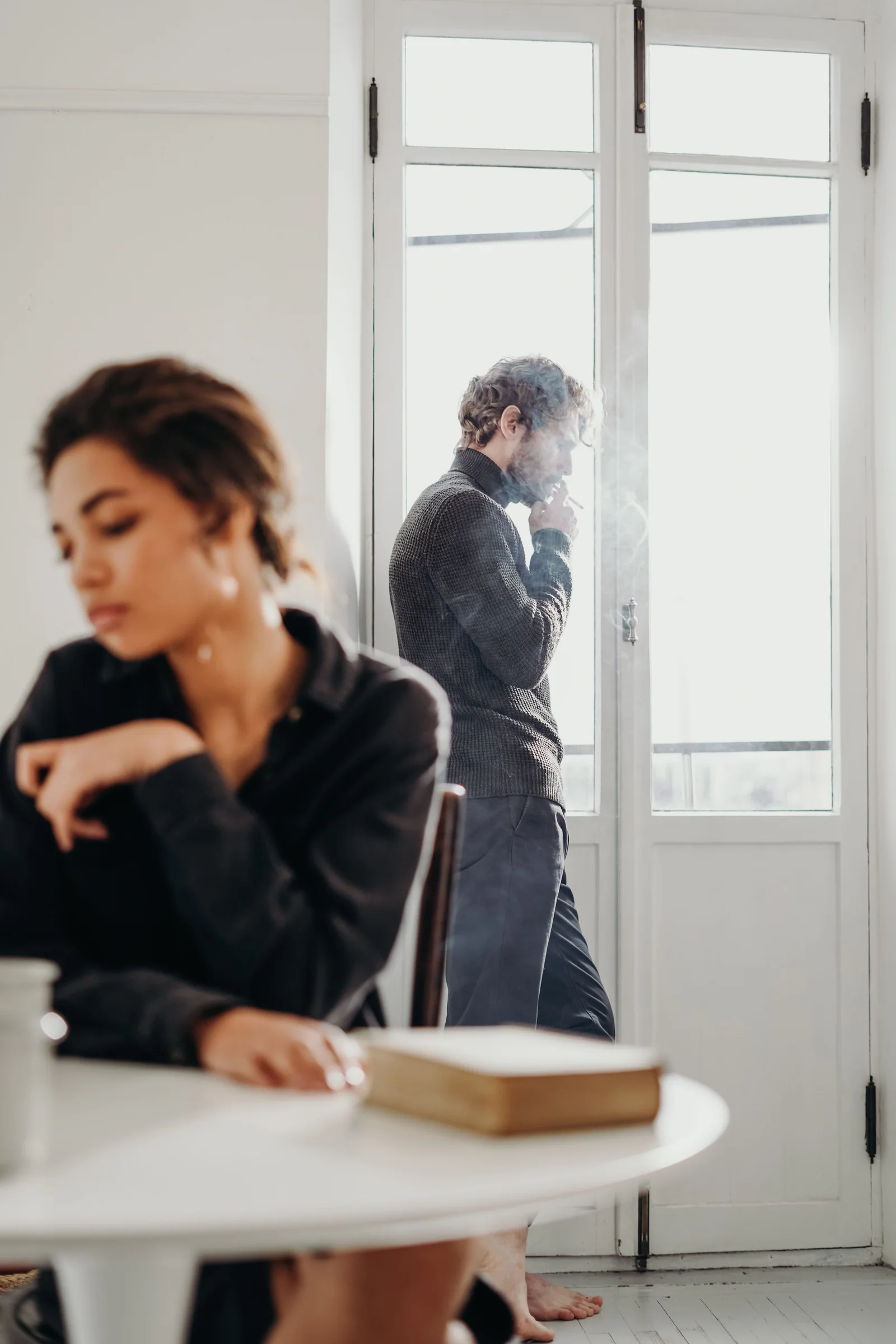 Donna seduta in primo piano e uomo che fuma vicino alla finestra